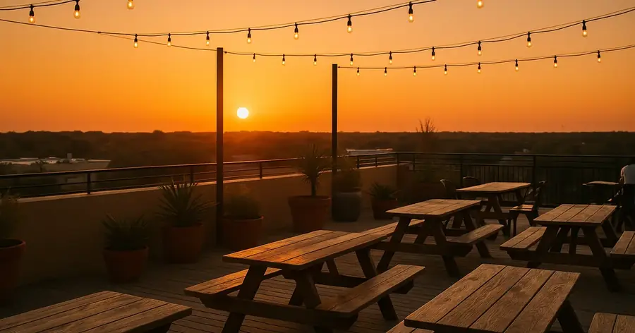 Rooftop taproom deck at sunset with weathered wood picnic tables, potted plants and cafe lights over a flat Texas treeline