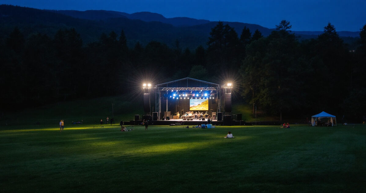 Outdoor amphitheater concert lawn at dusk with stage lights in a forested setting
